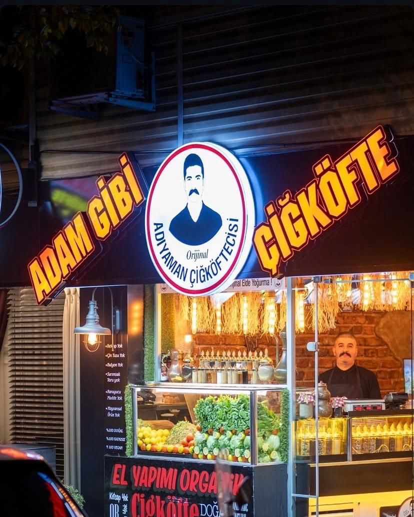 Adam Gibi Çiğköfte'nin gece cephesi: yanan ADAM GİBİ ve ÇİĞKÖFTE tabelaları, vitrinde taze yeşillikler ve tezgâhta usta.